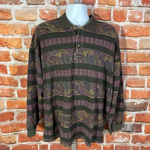 May include: A long-sleeved polo shirt with a paisley and striped pattern in shades of green, purple, and gold. The shirt has a collared neckline and button closure. The shirt is displayed on a white mannequin.