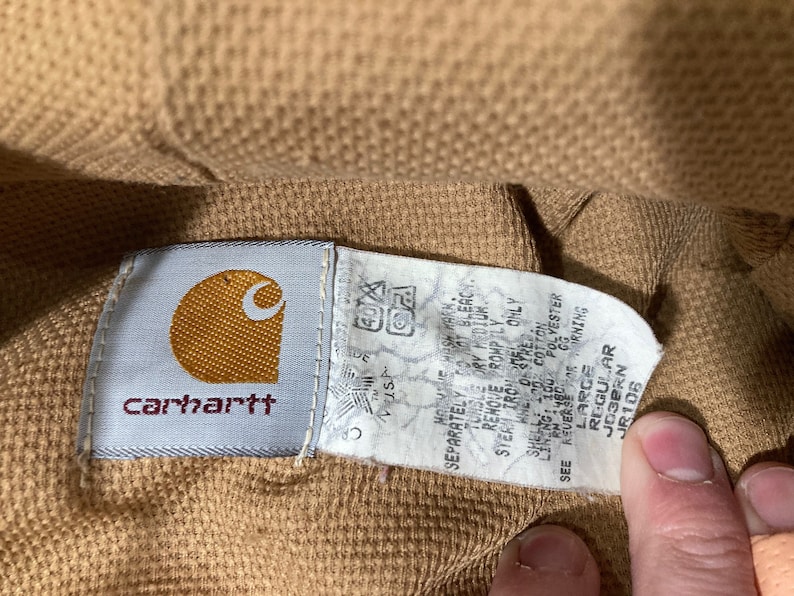 May include: A brown Carhartt workwear jacket with a white tag that reads "Must 40 Separately to not bleach Tumble dry low Remove promptly from dryer Only 100% Cotton 100% Polyester GG Shell Lining See care instructions", and a brown tag that reads "Large Regular Josern JR106".