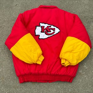 May include: A red puffer jacket with yellow sleeves and a ribbed red hem. The back features a large white arrowhead-shaped patch with a red and black KC logo in the center. The jacket has a high collar and long sleeves with elasticized cuffs.