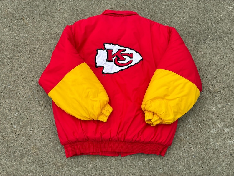 May include: A red puffer jacket with yellow sleeves and a ribbed red hem. The back features a large white arrowhead-shaped patch with a red and black KC logo in the center. The jacket has a high collar and long sleeves with elasticized cuffs.