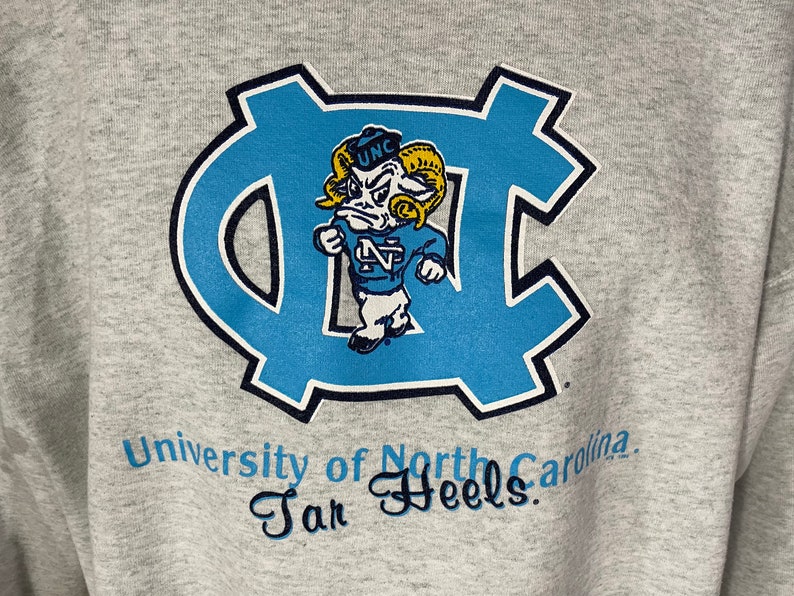Puede incluir: Sudadera gris con un logotipo azul y blanco de la Universidad de Carolina del Norte Tar Heels. El logotipo presenta un carnero de dibujos animados que lleva un sombrero que dice "UNC".