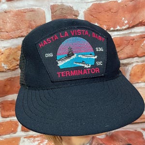 May include: Black trucker hat with a curved brim and mesh back. The front features a patch with the words "Hasta La Vista, Baby" and "Terminator" above an image of boats and a ship.