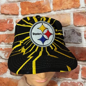 May include: A black baseball cap featuring a circular embroidered patch with the Pittsburgh Steelers logo in yellow, red, and blue. The cap is decorated with yellow lightning bolt embroidery radiating from the center patch across the crown and brim.
