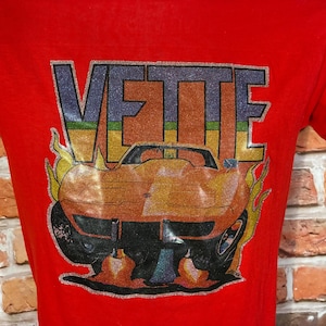 May include: A bright red t-shirt featuring a graphic print of an orange sports car with flames. The text VETTE is printed in large, glittery block letters above the car. The design includes a horizontal rainbow stripe and yellow flame accents.