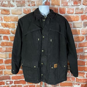 May include: Black Carhartt jacket with a button-up front, two chest pockets, and two lower pockets. The jacket has a collar and a Carhartt logo patch on the lower right pocket. The jacket is hanging on a white hanger.