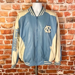 May include: Light blue and cream-colored varsity jacket with the "Tar Heels" text on the left sleeve and a logo on the chest. The jacket has a zip-up front, striped cuffs, and a collar. The jacket is displayed on a mannequin.