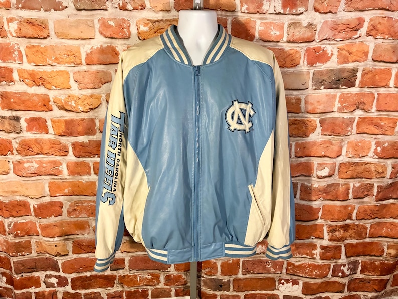 May include: Light blue and cream-colored varsity jacket with the "Tar Heels" text on the left sleeve and a logo on the chest. The jacket has a zip-up front, striped cuffs, and a collar. The jacket is displayed on a mannequin.