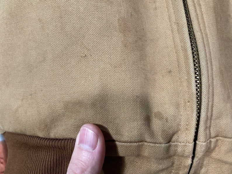 May include: Close-up of a brown canvas jacket with a zipper. The zipper is metal and has a silver finish.