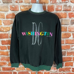 Puede incluir: Sudadera negra con cuello redondo verde y blanco. La parte delantera de la sudadera tiene un gran gráfico de las letras "DC" con la palabra "WASHINGTON" en letras de colores debajo.