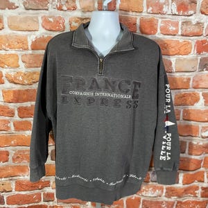 May include: A gray sweatshirt with a quarter-zip collar. The front features the word "FRANCE" above "COMPAGNIE INTERNATIONALE EXPRESS". The right sleeve has "POUR LA VILLE" printed on it. The bottom hem has text in French.