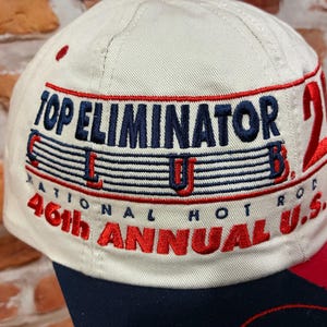 Op de afbeelding: Een beige honkbalpet met geborduurde tekst die Top Eliminator Club, National Hot Rod en 46th Annual U.S. Nationals leest. Het borduurwerk gebruikt marineblauw en rood garen, met een groot rood cijfer 21 zichtbaar op het zijpaneel.