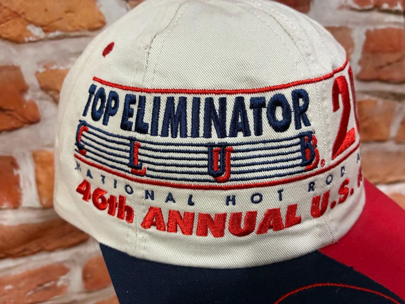 Op de afbeelding: Een beige honkbalpet met geborduurde tekst die Top Eliminator Club, National Hot Rod en 46th Annual U.S. Nationals leest. Het borduurwerk gebruikt marineblauw en rood garen, met een groot rood cijfer 21 zichtbaar op het zijpaneel.