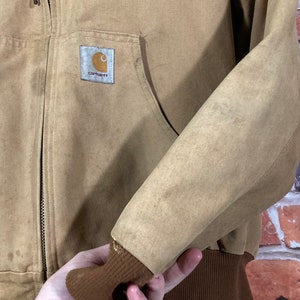 May include: A brown Carhartt work jacket with a zipper closure and a brown ribbed knit cuff. The jacket has a Carhartt logo patch on the left chest.