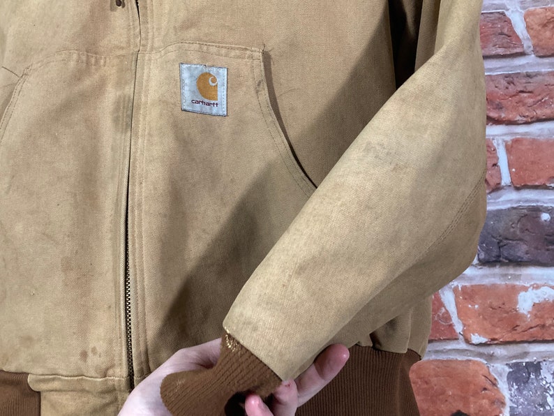 May include: A brown Carhartt work jacket with a zipper closure and a brown ribbed knit cuff. The jacket has a Carhartt logo patch on the left chest.
