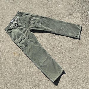 Op de afbeelding: Olijfgroene corduroy broek met twee cargozakken op de benen. De broek heeft een knoop- en ritssluiting en riemlussen. De stof heeft een zachte, getextureerde uitstraling. Deze broek is een casual stijl.