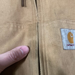 May include: A tan Carhartt work jacket with a zipper closure and a Carhartt logo patch on the right side. The jacket has a pocket on the left side.