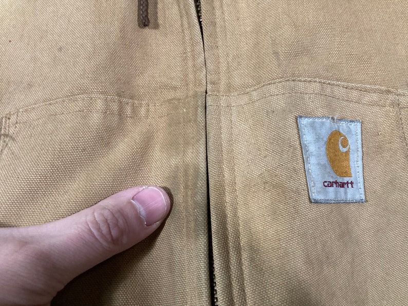 May include: A tan Carhartt work jacket with a zipper closure and a Carhartt logo patch on the right side. The jacket has a pocket on the left side.
