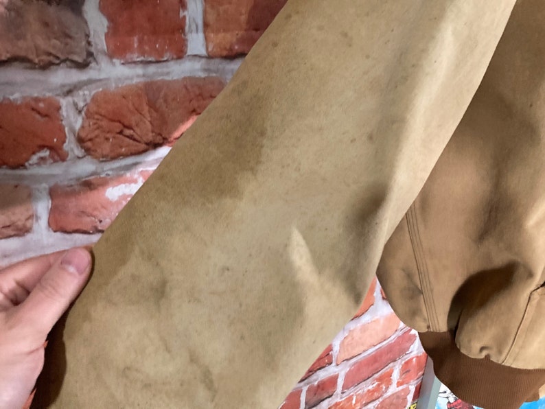 May include: Close-up of the tan-colored sleeve of a jacket. The fabric has a worn look with several brown stains.