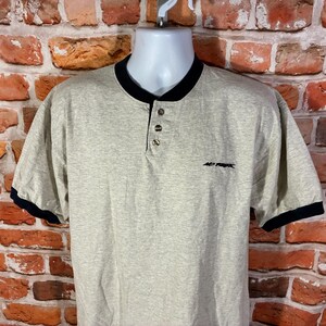 May include: A short-sleeve henley shirt in a heathered beige color with a black ribbed collar and black sleeve cuffs. The shirt features a three-button placket at the neckline and the text "NO FEAR" embroidered in black on the upper left chest.