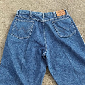 May include: Blue denim jeans with two back pockets and a brown leather patch. The jeans have a classic design with a straight leg and a button and zipper closure. The Wrangler logo is visible on the patch.