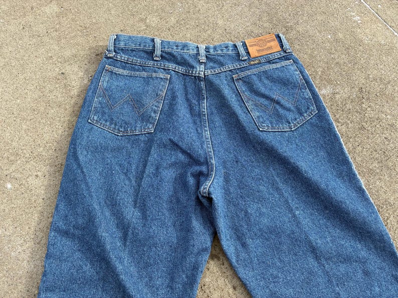 May include: Blue denim jeans with two back pockets and a brown leather patch. The jeans have a classic design with a straight leg and a button and zipper closure. The Wrangler logo is visible on the patch.