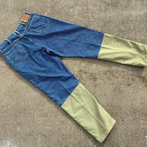 May include: Blue denim jeans with olive green lower legs. The jeans have a classic five-pocket design, a brown leather patch, and a button and zipper closure. The pants are laid flat on a concrete surface.