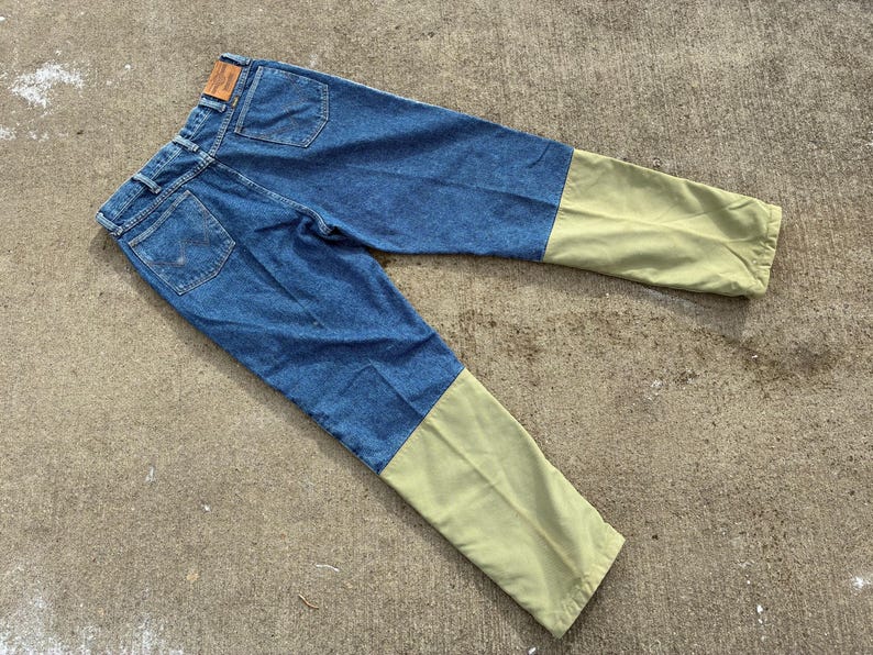 May include: Blue denim jeans with olive green lower legs. The jeans have a classic five-pocket design, a brown leather patch, and a button and zipper closure. The pants are laid flat on a concrete surface.