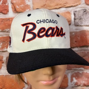 May include: Cream-colored baseball cap with a black brim. The cap features the text "CHICAGO Bears" in navy blue and red stitching. A logo is visible on the side of the cap. The cap is set against a brick wall.