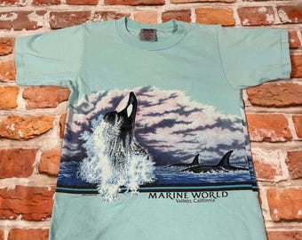 May include: A light blue t-shirt with a graphic print of an orca whale jumping out of the water. The graphic also includes a cloudy sky and the text "MARINE WORLD Vallejo, California".