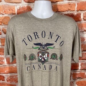 May include: A gray striped t-shirt with a graphic print of the word "TORONTO" in black lettering, a stylized eagle with a shield, and the word "CANADA" in black lettering.