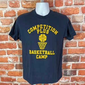 May include: A navy blue t-shirt with yellow text that reads "Competition Plus Basketball Camp" and a basketball and hoop graphic.