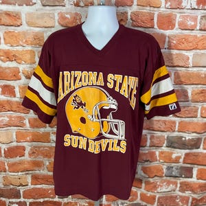 May include: Maroon v-neck t-shirt with gold and white striped sleeves. The shirt features a large graphic of a gold football helmet with the words "ARIZONA STATE SUN DEVILS" in gold lettering.