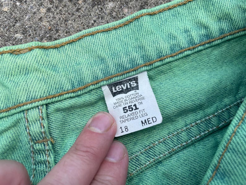May include: Close-up of a pair of green Levi's 551 jeans. The tag reads "551 Relaxed Fit Tapered Leg" and size 18. The jeans are made of 100% cotton and made in Canada. The stitching is visible.