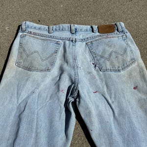 Peut inclure: Jean clair d&eacute;lav&eacute; avec des d&eacute;tails us&eacute;s, notamment des trous et des &eacute;claboussures de peinture. Le jean pr&eacute;sente une coupe classique &agrave; cinq poches, un patch en cuir et le logo Wrangler sur les poches arri&egrave;re. Aspect vintage.