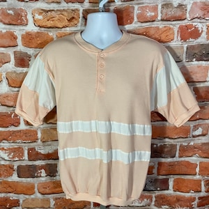 Peut inclure: Un t-shirt à manches courtes de couleur pêche avec des rayures horizontales blanches et une patte de boutonnage à un bouton. Le t-shirt est en tissu gaufré.