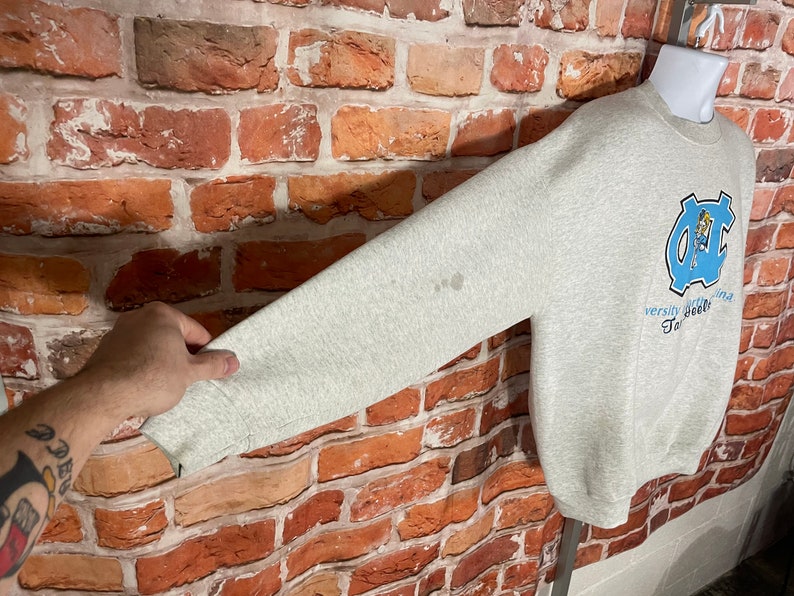 Puede incluir: Sudadera gris con un logotipo azul y blanco de la Universidad de Carolina del Norte Tar Heels y el texto "Varsity Carolina Tar Heels" en la parte delantera.