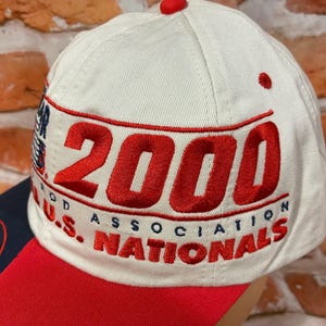 Op de afbeelding: Een cr&egrave;mekleurige baseballpet met rode geborduurde tekst die 2000 U.S. NATIONALS en ROD ASSOCIATION leest. De pet heeft een rode klep en een kleine rode knoop aan de bovenkant. Het borduurwerk is opvallend en duidelijk op de lichte stof.