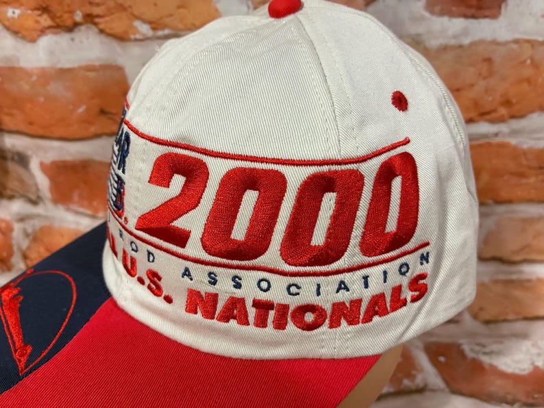 Op de afbeelding: Een cr&egrave;mekleurige baseballpet met rode geborduurde tekst die 2000 U.S. NATIONALS en ROD ASSOCIATION leest. De pet heeft een rode klep en een kleine rode knoop aan de bovenkant. Het borduurwerk is opvallend en duidelijk op de lichte stof.