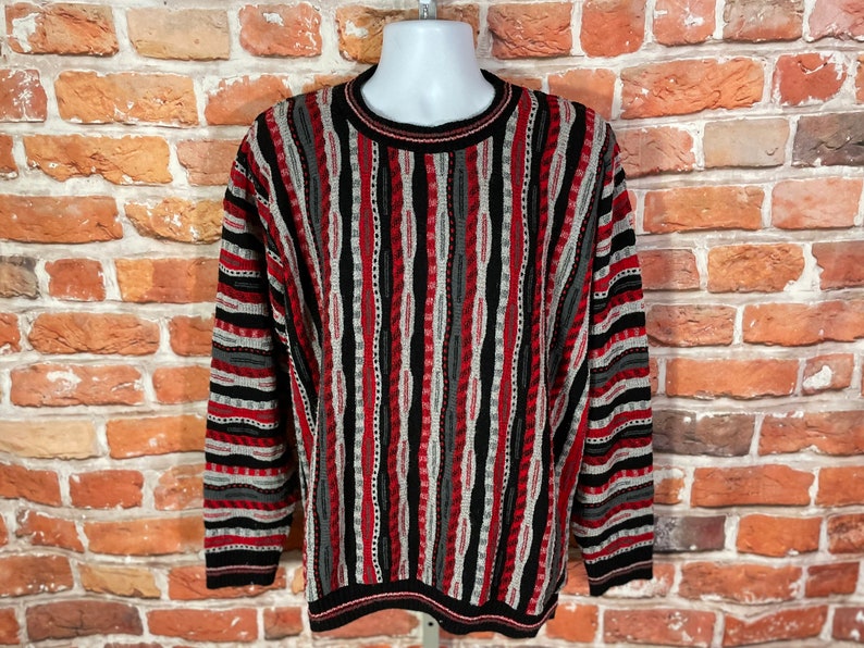 Peut inclure: Un pull ray&eacute; avec un motif noir, rouge et gris. Le pull a un col rond et des manches longues.