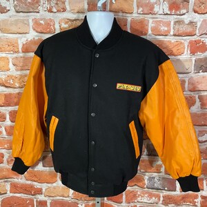 May include: Black and orange Pac-Man varsity jacket. The jacket features a black body with orange sleeves and a small Pac-Man logo on the chest. It has a button-up front and black ribbed cuffs and hem.