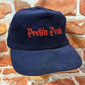 May include: Navy blue corduroy baseball cap with the text "Peek'n Peak" embroidered in red. The cap has a curved brim and a structured crown. The hat is displayed against a brick wall background.