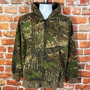 May include: A camouflage zip-up hooded jacket with a brown and green leaf pattern. The jacket has a full zipper, a hood, and brown cuffs and hem. The jacket is displayed on a white mannequin against a brick wall.