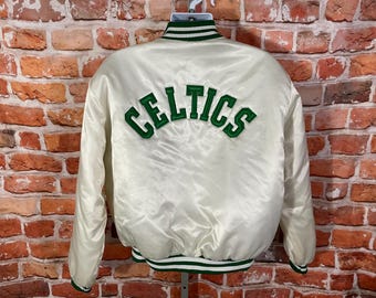 May include: Cream-colored satin jacket with green and white striped trim. The word "CELTICS" is embroidered in green across the back. The jacket is hanging on a white hanger against a brick wall.
