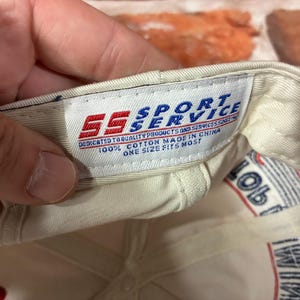 Op de afbeelding: De binnenkant van een gebroken witte honkbalpet met een wit stoffen label met de rood-blauwe tekst SS SPORT SERVICE, DEDICATED TO QUALITY PRODUCTS AND SERVICES SINCE 1982, 100% COTTON MADE IN CHINA, ONE SIZE FITS MOST.