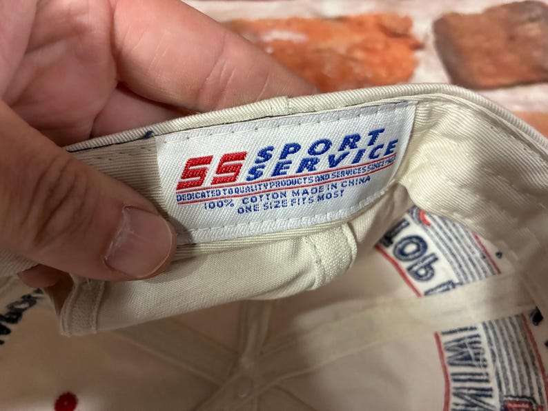 Op de afbeelding: De binnenkant van een gebroken witte honkbalpet met een wit stoffen label met de rood-blauwe tekst SS SPORT SERVICE, DEDICATED TO QUALITY PRODUCTS AND SERVICES SINCE 1982, 100% COTTON MADE IN CHINA, ONE SIZE FITS MOST.