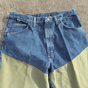 May include: Blue denim jeans with olive green panels on the lower front legs. The jeans have a classic five-pocket design, a button and zipper closure, and belt loops. The fabric appears to be a medium weight.