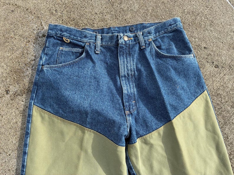 May include: Blue denim jeans with olive green panels on the lower front legs. The jeans have a classic five-pocket design, a button and zipper closure, and belt loops. The fabric appears to be a medium weight.