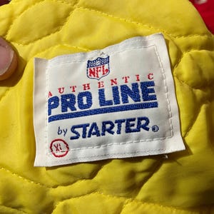 May include: A close-up of a white tag with the NFL logo, the words "AUTHENTIC PRO LINE by STARTER" in blue and red, and an XL size indicator. The tag is on a yellow quilted fabric.