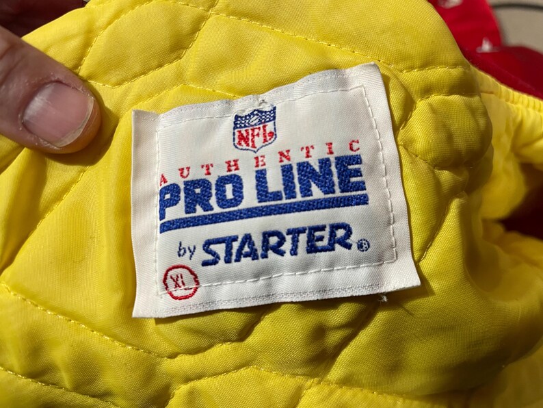 May include: A close-up of a white tag with the NFL logo, the words "AUTHENTIC PRO LINE by STARTER" in blue and red, and an XL size indicator. The tag is on a yellow quilted fabric.