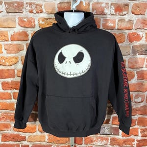 May include: Black hooded sweatshirt featuring a white Jack Skellington face on the front. The sleeve has the text "THE NIGHTMARE BEFORE CHRISTMAS" in red. The sweatshirt has a front pocket and a drawstring hood.